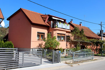 Fototapeta na wymiar Renovated small suburban family house with new dark pink facade and top balcony filled with flowers surrounded with garden plants and metal fence next to other houses and paved street on warm sunny