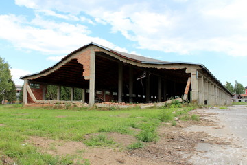 Obraz premium Wide large hangar building with missing destroyed red brick support walls and unusual bent roof surrounded with paved area mixed with grass and trees at abandoned military complex on cloudy blue sky b