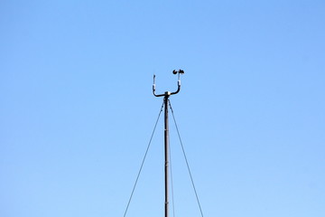 Wind speed and direction instruments mounted on top of tall metal pole held together with three strong wires on clear blue sky background