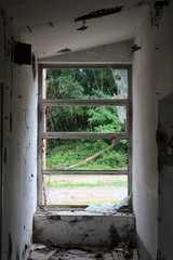 Wooden frame windows with shattered glass surrounded with white dilapidated cracked walls and garbage overlooking dense trees and grass at abandoned military complex