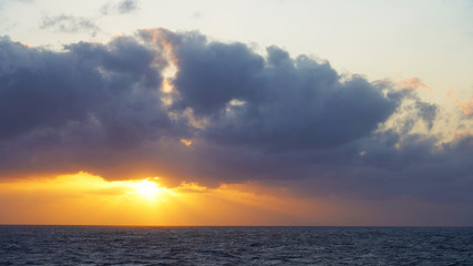 Fototapeta premium Orange-yellow sunset over the ocean. bright sunshine from beneath the clouds over the sea. rainy clouds sunrise 