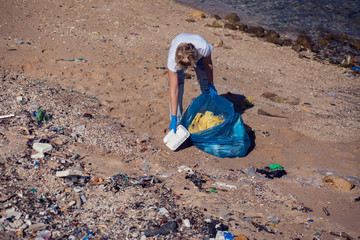 Obraz premium Woman volunteer with big blue bag collecting garbage on beach. Environmental pollution concept