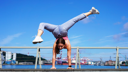 a young athletic girl equilibrist performs a stand on one arm on a sunny day. circus performer on the street. girl gymnast in life style clothes on the observation deck in the port. flexibility girl