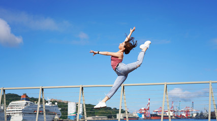 beautiful sporty girl in life style clothes jumping dance pose on the observation deck on a sunny day. circus gymnast in the port in denim casual wear. acrobatic twine jumping. gymnastic split in jump