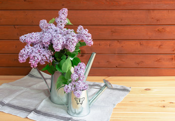 Two metal watering cans with branches of lilac on wooden table. View with copy space.