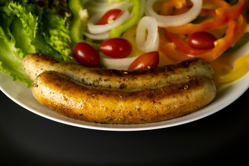 Two long grilled bratwurst sausage with mix vegetables salad in the white plate isolated on black background