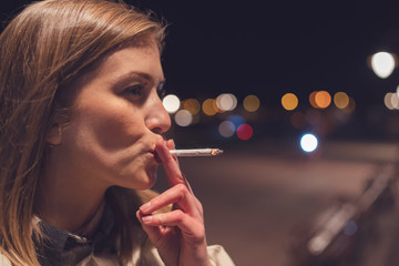 Obraz premium Close up portrait of young caucasian woman smoker on the street at night with cigarette smoking in the city outdoor smoke enjoining