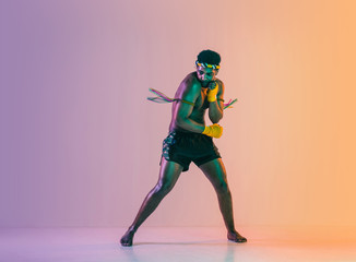 Fototapeta na wymiar Muay thai. Young man exercising thai boxing on gradient background in neon light. Fighter practicing, training in martial arts in action, motion. Healthy lifestyle, sport, asian culture concept.