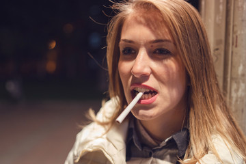 Fototapeta premium Close up portrait of young caucasian woman on the street at night holding cigarette in her mouth with teeth looking mean and furious mad