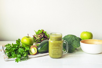 Detox green smoothie in a glass jar with ingredients: apples, kiwi, avocado, lettuce, broccoli, dill and parsley on white wooden table. Healthy raw vegan food concept.