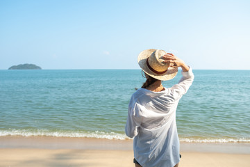 Back view or behind view of beautiful, young woman wearing white shirt and straw hat standing on beach with sea and sky background