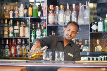 Portrait of professional african bartender man pours whiskey to the client of the hotel bar. The...