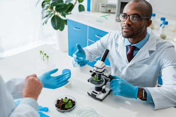 african american biologist sitting at table and looking at his colleague