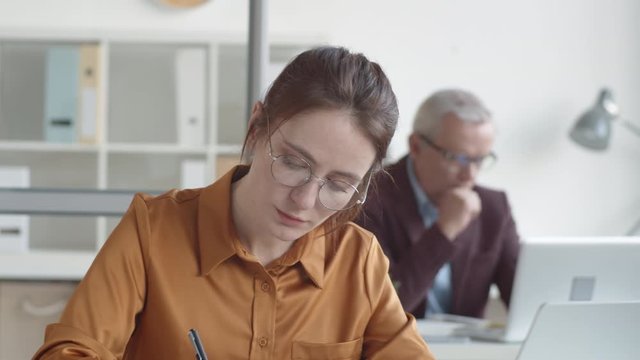 Tilting chest-up shot of nervous young Caucasian woman sitting at desk in office, looking at laptop screen and writing in journal, and middle-aged male superior in glasses working in background