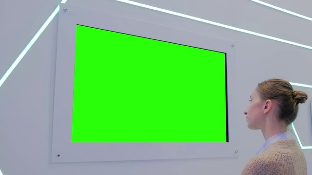 Green screen, mock up, future, copyspace, template, chroma key, technology concept. Woman looking at blank digital interactive green display wall at exhibition or museum with futuristic scifi interior