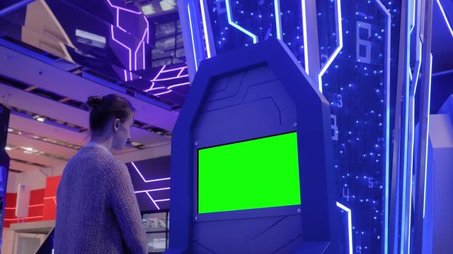 Woman looking at blank digital interactive green display kiosk at exhibition or museum with futuristic sci-fi interior. Mock up, copyspace, template, chroma key, green screen, technology concept