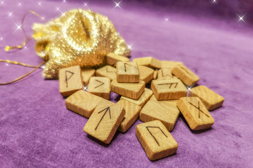 A number of Scandinavian runes made on wooden boards lie on a purple velvet fabric next to a golden shiny bag, attracting the gifts of God. Fortune telling tool, concept for predicting the future.