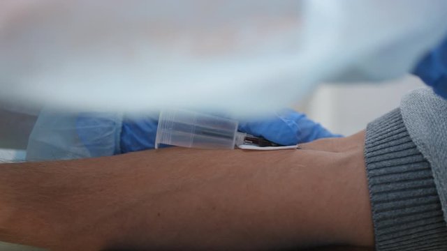 Tracking close up shot of hands of nurse in disposable gloves taking venous blood from arm of male patient, removing needle and pressing down on vessel with sterile gauze
