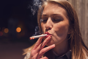 Close up portrait of young caucasian woman smoker on the street at night with cigarette smoking in...