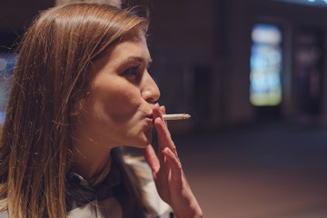 Close up portrait of young caucasian woman smoker on the street at night with cigarette smoking in...