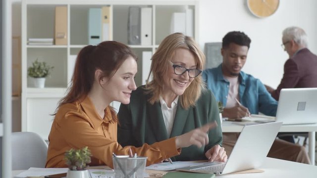 Waist-up shot of young Caucasian female office worker sitting at desk together with mature manager, showing her something on laptop screen and explaining while woman is watching and smiling