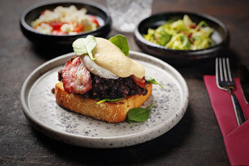 Toast with black pudding, poached egg, bacon and mayonnaise sauce. Food, ready dish on a dark...