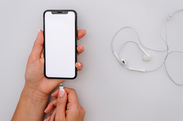 Woman hand holding the black smartphone with big blank screen and pluig in white headphones. Top view mockup
