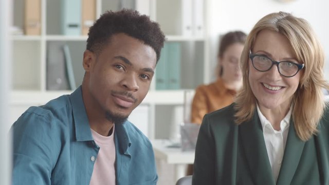 Chest-up shot of middle-aged Caucasian female HR manager sitting next to young Afro-American intern in office, talking, then looking at camera and smiling