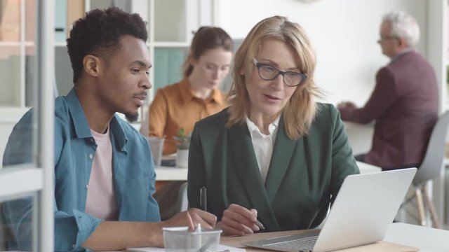 Waist-up shot of mature Caucasian female supervisor sitting at desk together with new male African American employee, explaining duties on laptop, while man is watching attentively and taking notes