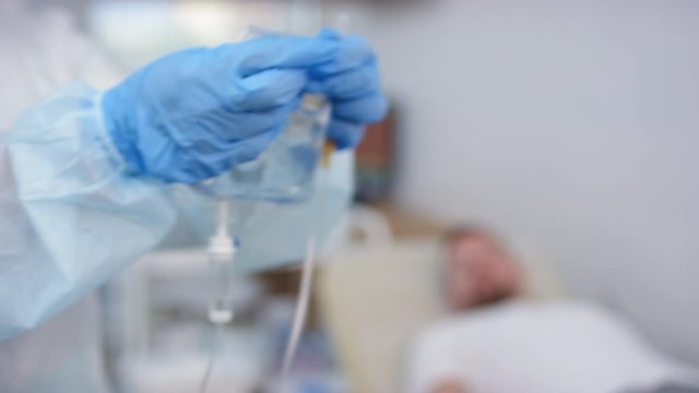 Selective focus shot of nurse in medical uniform hanging fluid bag on IV pole while preparing for giving intravenous therapy to male patient in clinic