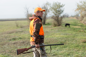 A man with a gun in his hands and an orange vest on a pheasant hunt in a wooded area in cloudy...