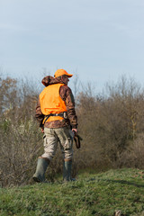 A man with a gun in his hands and an orange vest on a pheasant hunt in a wooded area in cloudy...
