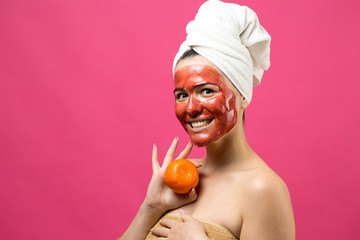 Beauty portrait of woman in white towel on head with red nourishing mask on face. Skincare cleansing eco organic cosmetic spa relax concept. A girl stands with her back holding an orange mandarin.