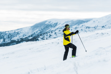 Naklejka na ściany i meble skier in helmet holding sticks and standing on slope in wintertime