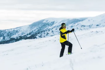 Fotobehang skier in helmet holding sticks and standing on slope in wintertime © LIGHTFIELD STUDIOS