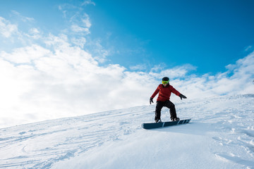 athletic snowboarder in helmet riding on slope against blue sky with clouds