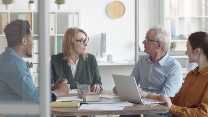 Waist-up shot of four diverse male and female colleagues sitting around table in office at team meeting, having lively discussion of work issues, making proposals and suggestions and laughing