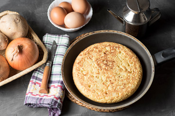 Spanish potato omelette, on a table, next to the necessary ingredients to be cooked and ready to be eaten