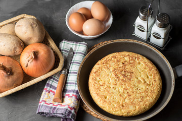 Spanish potato omelette, on a table, next to the necessary ingredients to be cooked and ready to be eaten