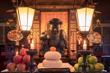 New year's mochi rice, clementine and apples offerings in front of a wooden statue depicting one of...