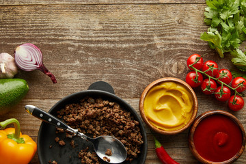 Top view of minced meat in frying pan with sauces and raw vegetables on wooden background