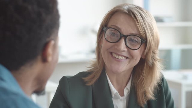 Close-up over-shoulder shot of smiling middle-aged Caucasian female HR director in glasses and business jacket talking to young Afro-American candidate during job interview in office