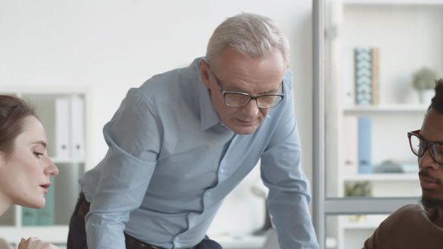 Chest-up handheld panning shot of multiethnic team sitting and senior Caucasian male executive standing at table during business meeting and discussing sales data on chart