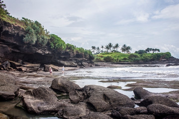 Two tourists wandering in rocky Tanah Lot beach in low tide. Palm Trees and dramatic cloudy sky in the background. Bali