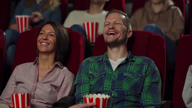 Tracking left medium shot of middle aged couple sitting on comfortable seats in cinema, eating popcorn, watching comedy movie and laughing