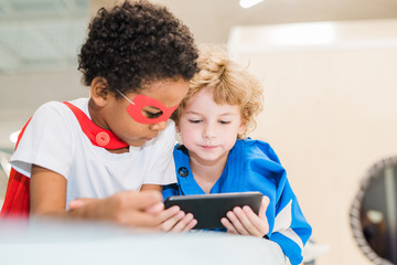 Two little intercultural friendly boys in costumes of superman watching video