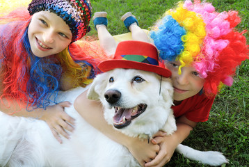 Small children dressed in elegant festive fun wigs with their dog and play with it in nature in the summer.