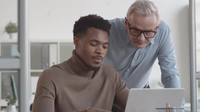 Medium shot of senior Caucasian male manager approaching young African American associate sitting and working on laptop at desk, asking him about work on project, man giving boss update and laughing