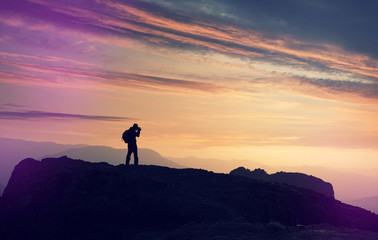 Silhouette of photographer, tourist on the edge of cliff at sunrise. Mountain valley during bright sunset. Beautiful natural landscape in the summer time. Man on top of mountain. Conceptual scene.