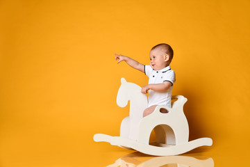 Playful infant baby boy toddler is riding his wooden horse for children and pointing his finger at free copy space